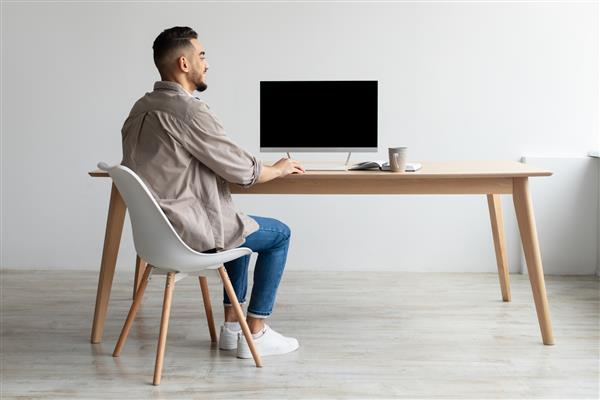 نمای پشتی مرد عرب خندان شاد که به رایانه شخصی با صفحه سیاه خالی برای الگوی ماکت نشسته روی میز در دفتر خانه فضای کپی رایگان نگاه می کند مردم فناوری مفهوم کار از راه دور