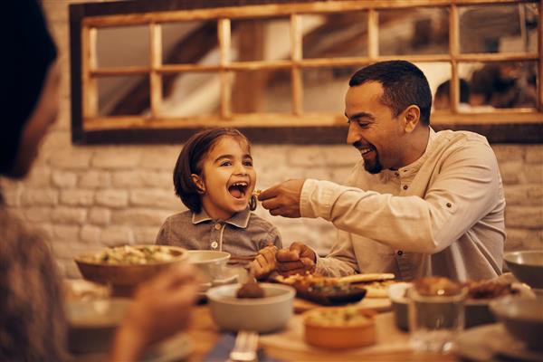 دختر کوچک مسلمان شاد در حالی که در ماه رمضان در خانه شام می خورد در حالی که پدر به او غذا می دهد سرگرم می شود
