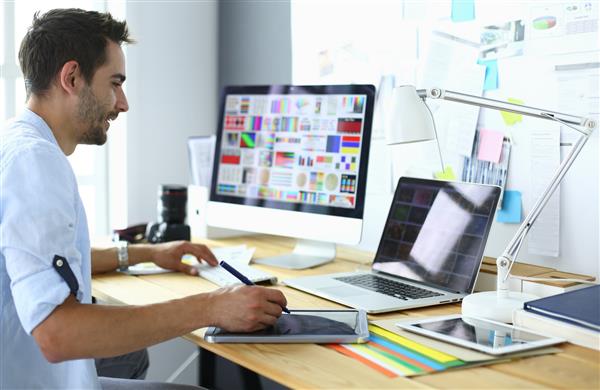 پرتره طراح جوانی که در استودیوی گرافیکی در مقابل لپ‌تاپ و کامپیوتر نشسته در حالی که آنلاین کار می‌کند