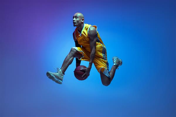 پرواز بسکتبالیست مرد خوش تیپ آفریقایی-آمریکایی در حال حرکت و اکشن با نور نئون روی دیوار آبی