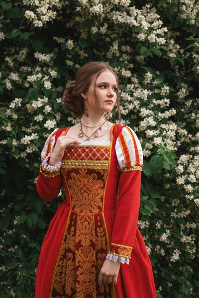 زن جوان زیبا با لباس قرون وسطایی قرمز در باغ با گل های سفید ایستاده است