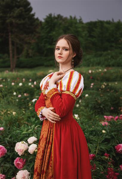 زن جوان زیبا با لباس قرون وسطایی قرمز با گل صد تومانی سفید در باغ ایستاده است