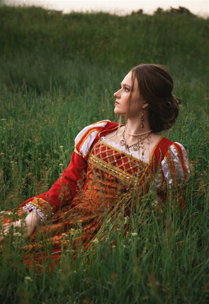 زن جوان زیبا با لباس قرون وسطایی قرمز بلند در چمن نشسته است