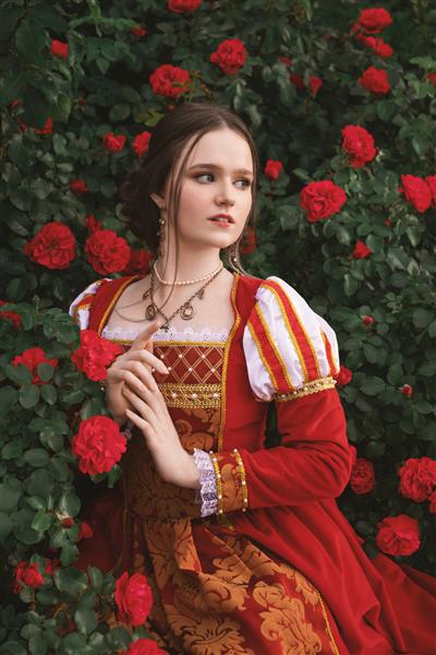 زن جوان زیبا با لباسی به سبک قرون وسطایی با گل رز قرمز در باغ نشسته است
