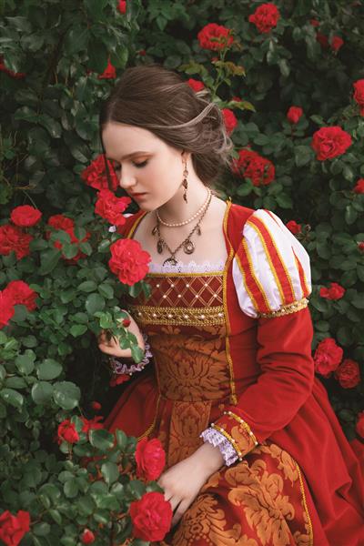 زن جوان زیبا با لباسی به سبک قرون وسطایی با گل رز قرمز در باغ نشسته است