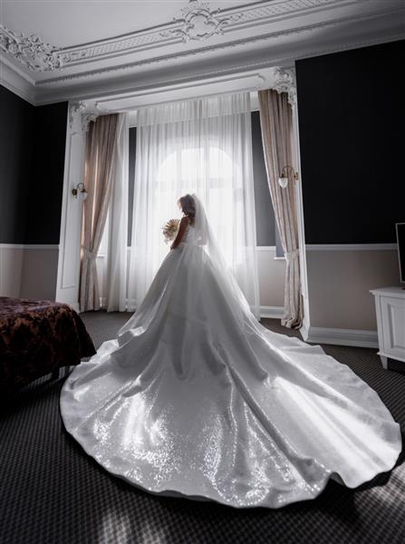 نمای پشت نامزد با لباس عروس پر پوش ایستاده در میان اتاق خالی و دسته گل در دست