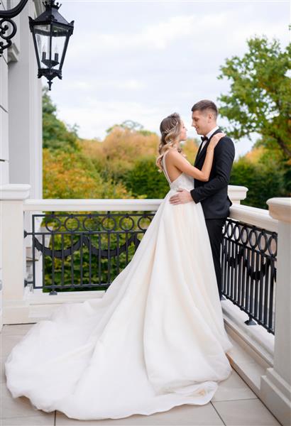 نمای کناری عروس جذاب با لباس عروس در آغوش داماد با کت و شلوار گران قیمت و دست در دست