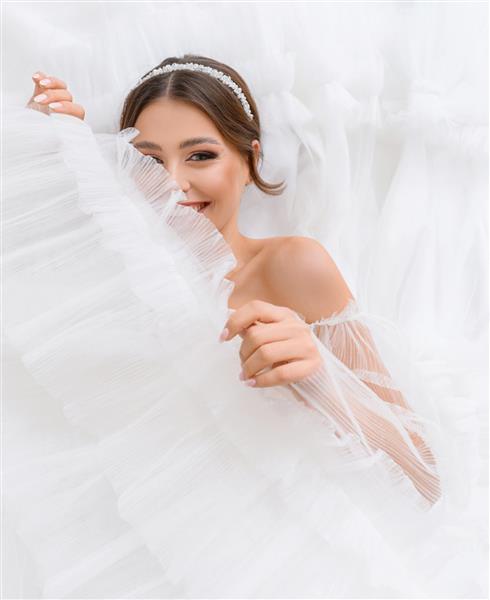 زن جوان دوست داشتنی شادی که روی لباس پف کرده سفید عروسیش دراز کشیده به دختر عروس خندان دوربین با آرایش برهنه و جواهرات ایده آل لباس عروس سالن عروسی عروس شایان ستایش نگاه می کند