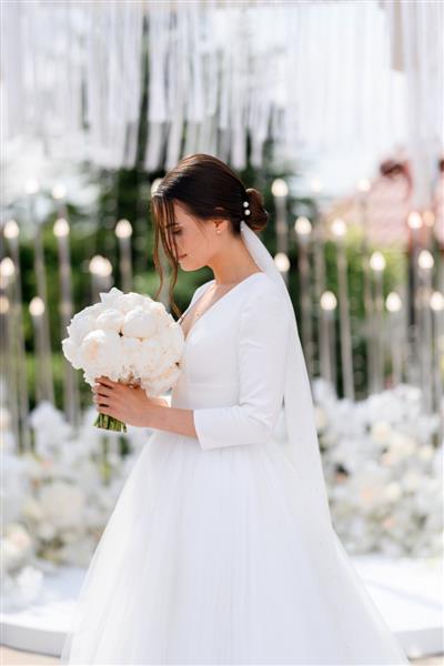 نمای جانبی زن سبزه عروس جذاب با محجبه و لباس عروسی زیبا که به دسته گل گل صد تومانی ایستاده است مراسم عروسی در محراب گلدار در فضای باز عروس زیبا
