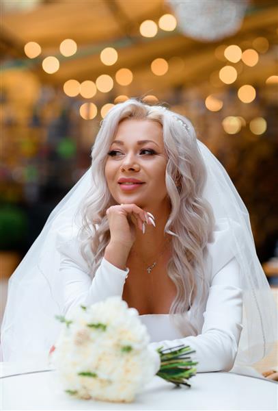 نمای جلوی عروس پر زرق و برق با مدل مو و حجاب در لباس عروس با یقه عمیق و اکسسوری های روی گردن که به سمت دور نگاه می کند و لبخند می زند