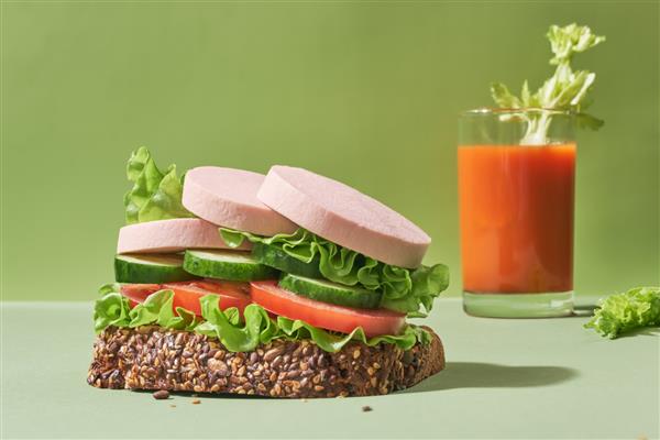 ساندویچ با سوسیس برش خورده و سبزیجات با یک لیوان آب میوه در زمینه سبز