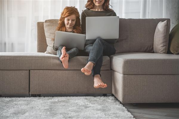 زاویه پایین کودک و مادرش با کامپیوتر در خانه نشسته است آنها در حالی که با هم در اینترنت گشت و گذار می کنند لپ تاپ ها را با لبخند و لذت در دست دارند