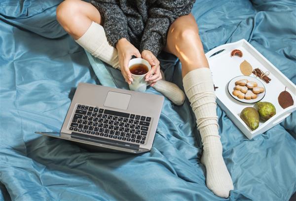 زن جوان در حال نوشیدن چای در خانه در رختخواب خود و چک کردن لپ تاپ خود نمای بالا