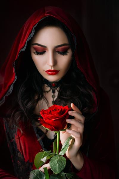 زن زیبای عرفانی در لباس گوتیک یک خون آشام قرون وسطایی در کاپوت یک دختر بالغ گل رز را در دستان خود نگه می دارد آرایش قرمز صورت زیبا لب های جذاب تصویر جشن هالووین