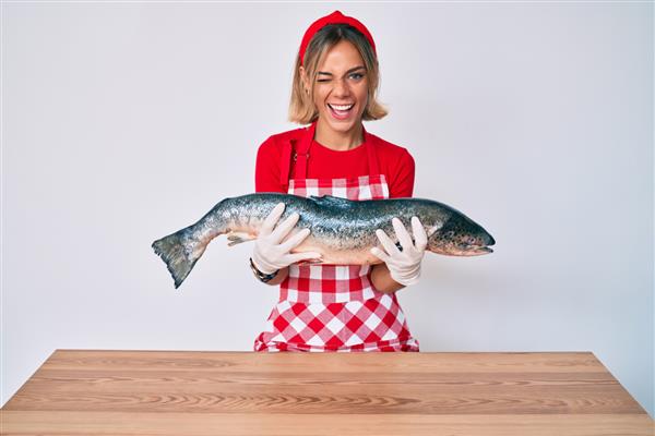 زن زیبای قفقازی ماهی فروش که ماهی قزل آلا خام تازه می فروشد و چشمک می زند و با حالتی جذاب چهره ای شاد و شاد به دوربین نگاه می کند