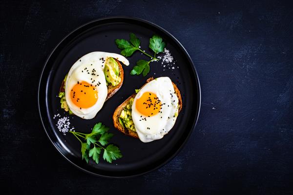 صبحانه قاره ای - تخم مرغ های آفتابی روی نان برشته شده با آووکادو در پس زمینه سیاه