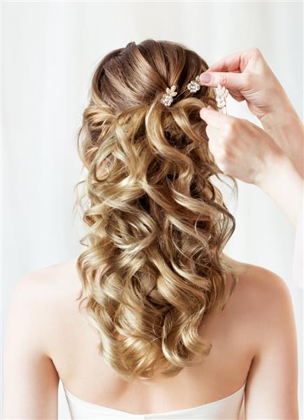 نمای پشتی مدل موی عروسی لوازم جانبی مدل موی عروس نمای عقب از نزدیک آرایشگاه در حال گذاشتن گیره موی کریستالی گل مدل موی شب موج دار روی پس زمینه سفید