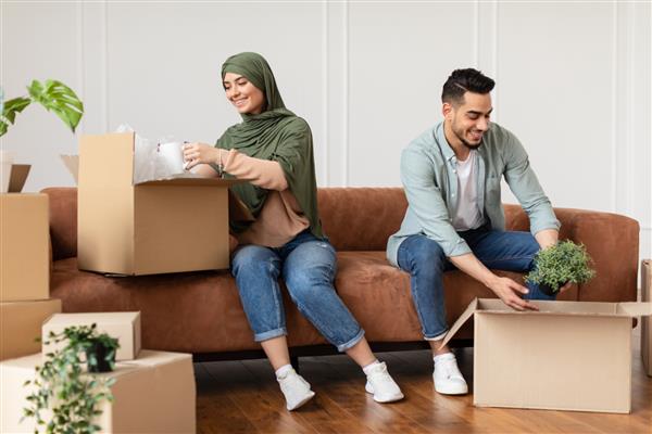 خرید املاک جابجایی خانه مفهوم جابجایی پرتره خانواده مسلمان هیجان زده در حال باز کردن جعبه های مقوایی نشستن با بسته بندی باز روی کاناپه در اتاق نشیمن مسکن جوانان