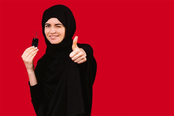 دختر مسلمان با حجاب مشکی کلید ماشین جدید را در دست دارد پلک می‌زند و جلوی دوربینی که کلاس را نشان می‌دهد ژست می‌گیرد انگشت شست روی پس‌زمینه قرمز با فضای کناری