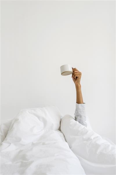مفهوم صبح زود بیدار شدن نمای عمودی زن با پوست تیره با لباس خواب روی تخت پیچیده شده در پتوی گرم و دنج دست را با فنجان قهوه بلند کنید دختری که لیوان را در برابر دیوار فضای کپی سفید نگه داشته است