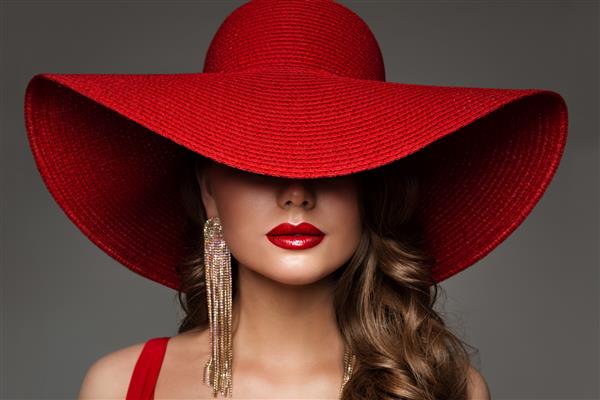 زن مد با کلاه با آرایش لب قرمز و گوشواره طلایی مدل زیبایی صورت پنهان شده توسط کلاه لبه پهن پرتره بانوی زیبا از نزدیک روی خاکستری