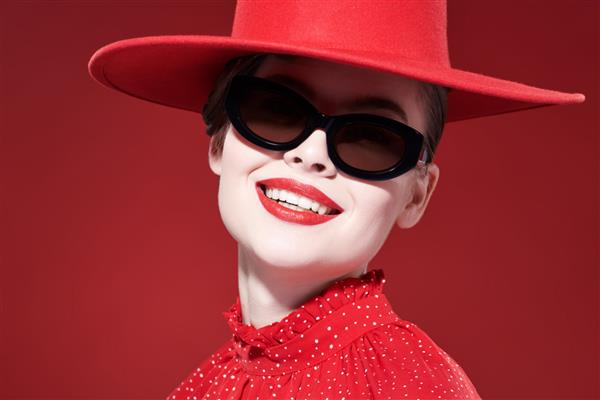 یک زن جوان شیک پوش جذاب با آرایش روشن با لباس قرمز کلاه قرمزی و عینک آفتابی شیک ژست می گیرد و لبخندی جذاب می زند پس زمینه قرمز زیبایی آرایش و لوازم آرایشی روش
