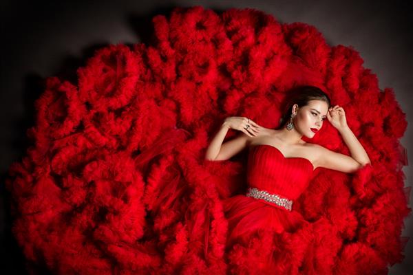 مدل مد لباس ابری کرکی قرمز دراز کشیده پرتره زن زیبا با جواهرات نقره بانوی زیبا با لباس مجلسی که از پس زمینه استودیو خاکستری نگاه می کند