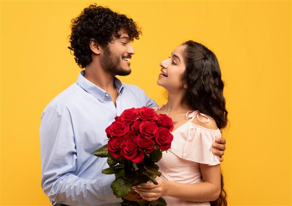 تعطیلات عاشقان زن و شوهر عاشق هندی که دسته گلی را در دست گرفته اند یکدیگر را در آغوش گرفته و روی پس زمینه زرد نگاه می کنند پسر جوانی که در روز ولنتاین دوست دخترش را با گل رز نوازش می کند