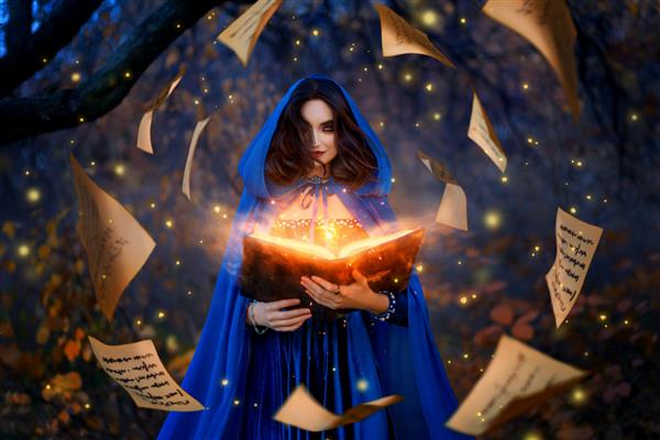 جادوگر زن فانتزی با کاپوت کتاب جادویی در دست دارد طلسم های نور نارنجی روشن باد برگه های پاییزی را پراکنده می کند دختر جادوگر لباس جادوگر با شنل آبی قرون وسطایی