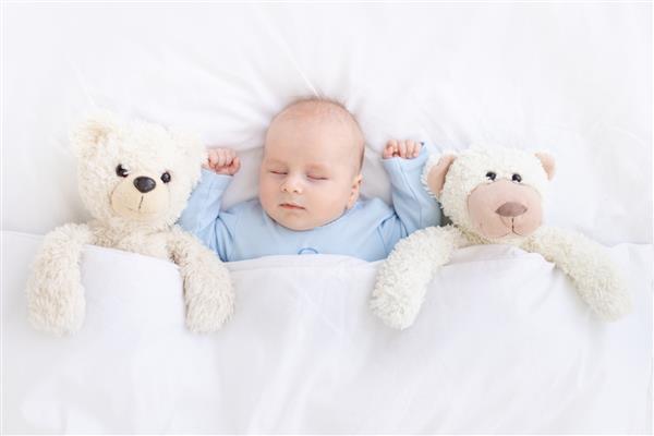 نوزاد پسر با یک خرس اسباب بازی نرم با لباس خواب آبی که دستانش را بالا گرفته روی تخت دراز کشیده به پشت می خوابد