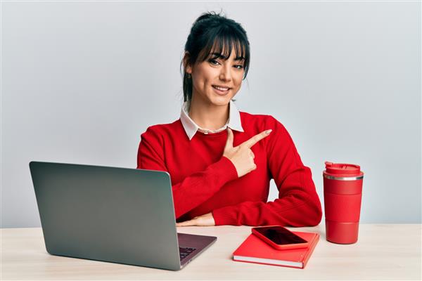 زن جوان سبزه با چتری که در دفتر کار می کند با لپ تاپ لبخند می زند که دست و انگشتش را به سمت بالا نشان می دهد