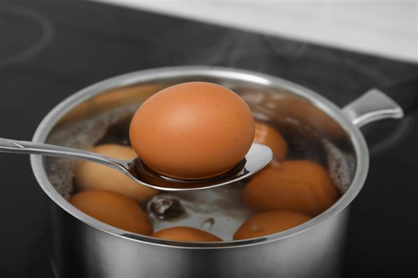قاشق با تخم مرغ آب پز بالای قابلمه روی اجاق برقی نمای نزدیک