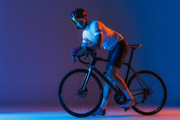 یک دوچرخه‌سوار مرد در حال دوچرخه‌سواری با شلوارک دوچرخه‌سواری و کلاه ایمنی جدا شده روی پس‌زمینه آبی تیره در نئون مفهوم زندگی فعال استراحت سفر انرژی ورزش فضای تبلیغاتی را کپی کنید