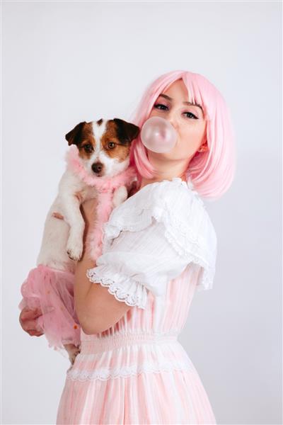 دختری با موهای صورتی و لباس صورتی حباب جویدنی را باد می‌کند و سگش را با لباس صورتی جدا شده روی پس‌زمینه سفید نگه می‌دارد دختری با موهای صورتی و کک و مک آدامس می زند