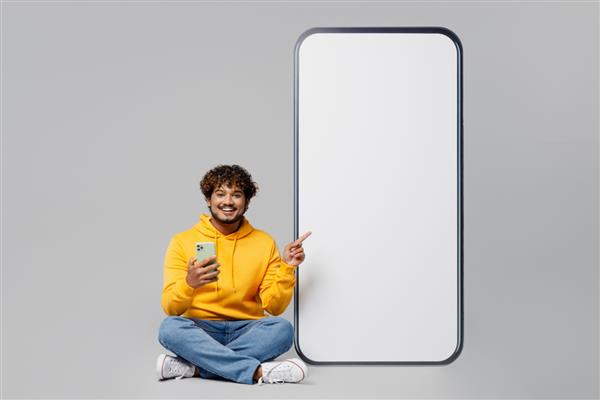 مرد جوان هندی با اندام کامل با هودی زرد معمولی و نشسته کنار تلفن همراه با صفحه‌نمایش خالی بزرگ با فضای نمایش فضای کاری از تلفن همراه جدا شده روی پرتره استودیو پس‌زمینه خاکستری ساده استفاده می‌کند