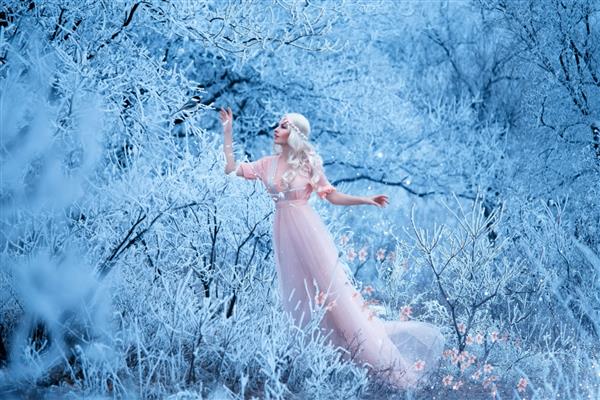 زن بلوند جوان شاهزاده خانم باند فرودگاه با لباس هلویی در جنگل آبی زمستانی راه می‌رود یخ زدگی و برف روی درختان داستان سفید برفی دختر بهار پرنده را از سرما گرم می کند گلها از او می ریزند