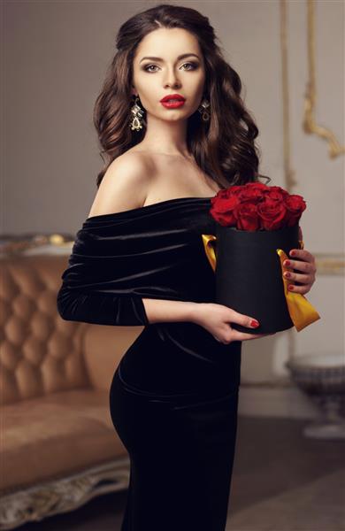 دختر زیبای جوان ایستاده و جعبه سیاه با گل رز قرمز را در دست دارد پرتره استودیویی به سبک مد ووگ از دختر جذاب با لباس مشکی شیک که در فضای داخلی مجلل ایستاده است