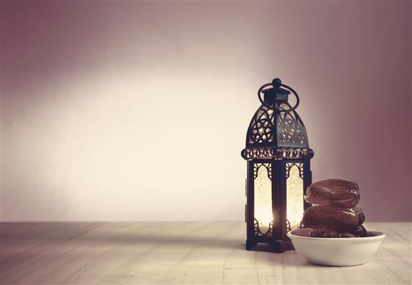 میوه‌های خرما روی ظرف سفید روی زمین چوبی کنار فانوس درخشان در زمینه رنگارنگ آماده سرو در شب کریمه رمضان