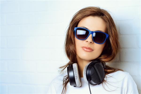 زن جوان مدرن در حال گوش دادن به موسیقی با هدفون در کنار دیوار آجری ایستاده است سبک جوانان