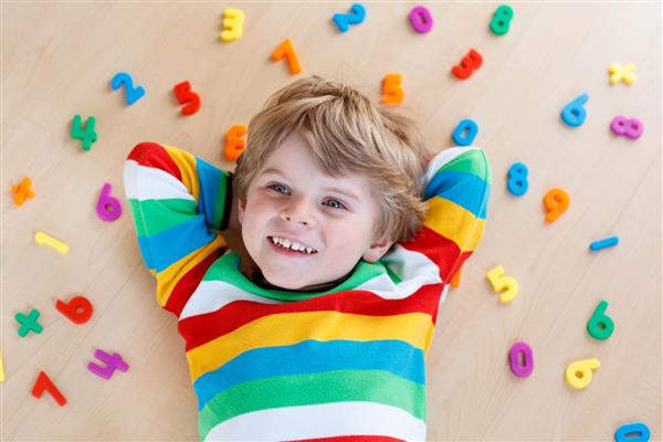 کودک نوپا بلوند کوچک در حال بازی با تعداد زیادی ارقام یا اعداد پلاستیکی رنگارنگ در داخل خانه پسر بچه ای که پیراهن رنگارنگ پوشیده و با یادگیری ریاضی سرگرم می شود