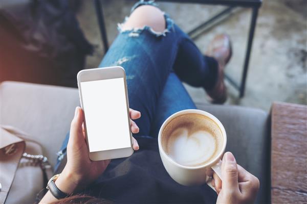 تصویر موکاپ از دستان زنی که تلفن همراه سفید را با صفحه خالی روی ران در دست گرفته و فنجان قهوه در کافه