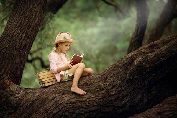 یک دختر بلوند کوچک با کلاه حصیری در حال خواندن کتاب روی درختی بزرگ کودکان و علم