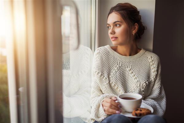 زن جوان زیبا در حال نوشیدن قهوه و نگاه کردن از پنجره در حالی که پشت طاقچه خانه نشسته است