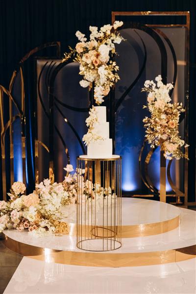 عروسی کیک سه طبقه سفید تزئین شده با گل روی میز طلایی در محوطه تزئین شده قرار دارد