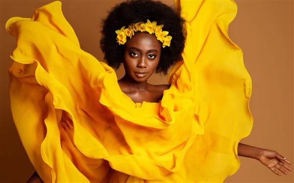 پرتره مد زن با پوست تیره در لباس پرنده زرد مدل آفریقایی زیبایی با اکلیل گل در مدل موی سیاه آفریقایی رقصنده با پارچه ابریشمی در حال بال زدن روی پس زمینه بژ استودیو