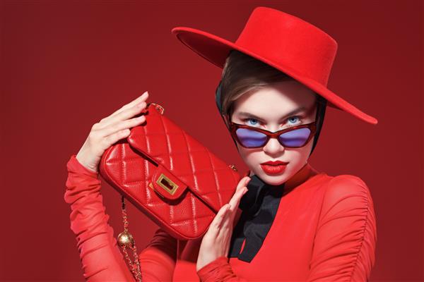 مد شات یک دختر زیبا و شیک با آرایش روشن و عینک با لباس های قرمز ظریف با یک کیف دستی قرمز شیک ژست می گیرد پس زمینه قرمز تجهیزات جانبی