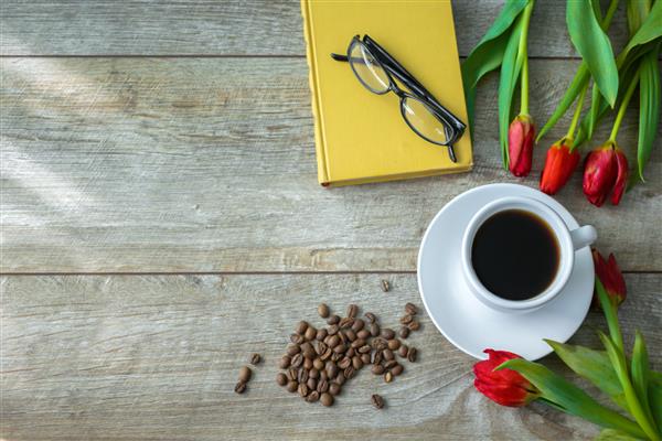 نمای بالای فنجان قهوه اسپرسو و دانه ها دسته گل لاله قرمز گل نوشیدنی داغ و کتاب روز مادر زنان مفهوم صبح بهار بنر تخت روی میز چوبی با فضای کپی