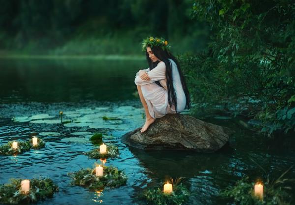 دختر اسلاوی روی سنگ در ساحل دریاچه نشسته است زن فانتزی پوره زانوها را در آغوش می گیرد موهای بلند مشکی تاج گل های علف گل ها روی آب شناورند شمع در حال سوختن درخت سبز جنگل غروب رودخانه آیین فال