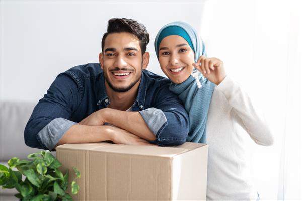 جاکلیدی در دستان زوج مسلمان خندان زن و شوهر خانواده خوشبخت خاورمیانه ای در حال نشان دادن کلید آپارتمانشان جشن رفتن به خانه جدید ایستادن در کنار جعبه های کاغذی نمای نزدیک