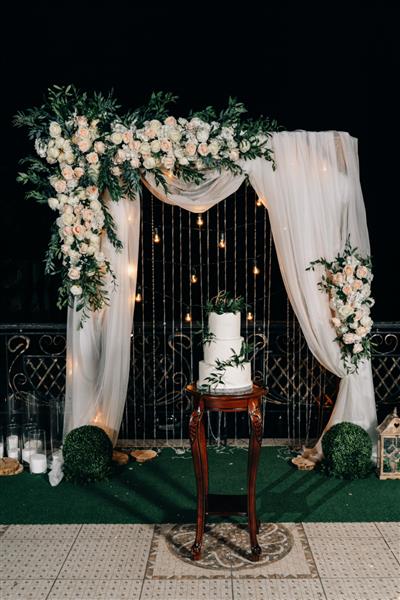 کیک عروسی سفید سه طبقه با نوشته LOVE خامه ماستیک سفید تزئین شده با گیاهان روی پایه چوبی